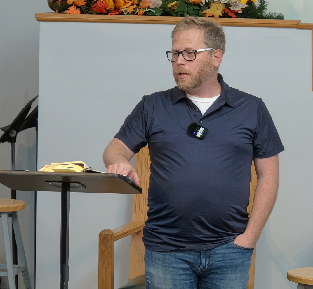 A man with a beard and glasses stands next to a lectern in a room decorated with autumn leaves, gesturing while speaking.