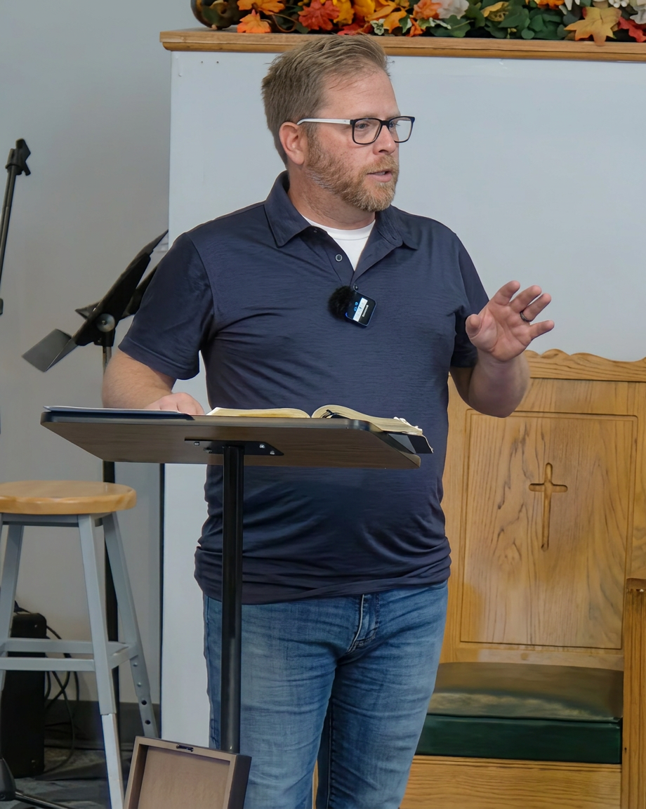 A man with glasses gestures while speaking at a lectern in a church setting, holding an open book, with wooden furnishings in the background.