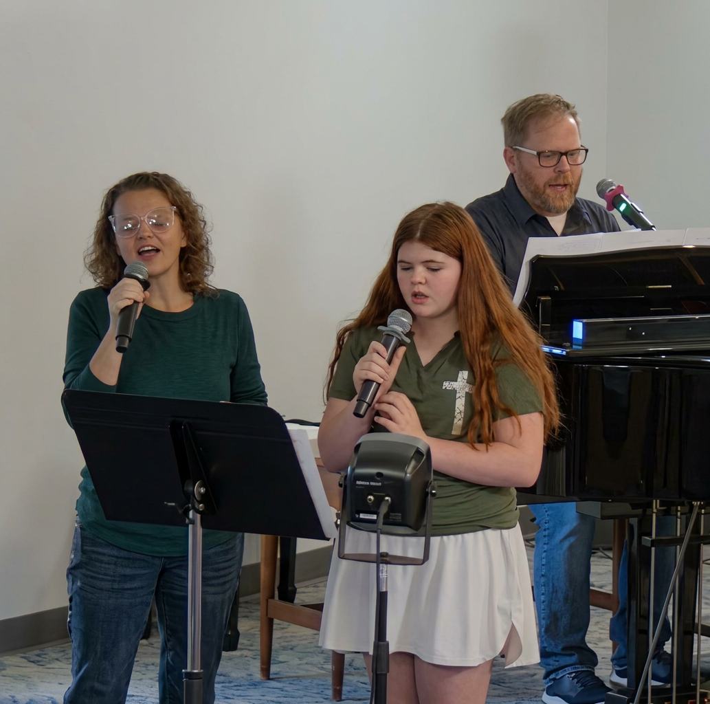 Two singers performing on stage with microphones, accompanied by a pianist. A woman on the left sings passionately, while a girl in the middle focuses on her performance. A man at a piano in the background provides musical support.
