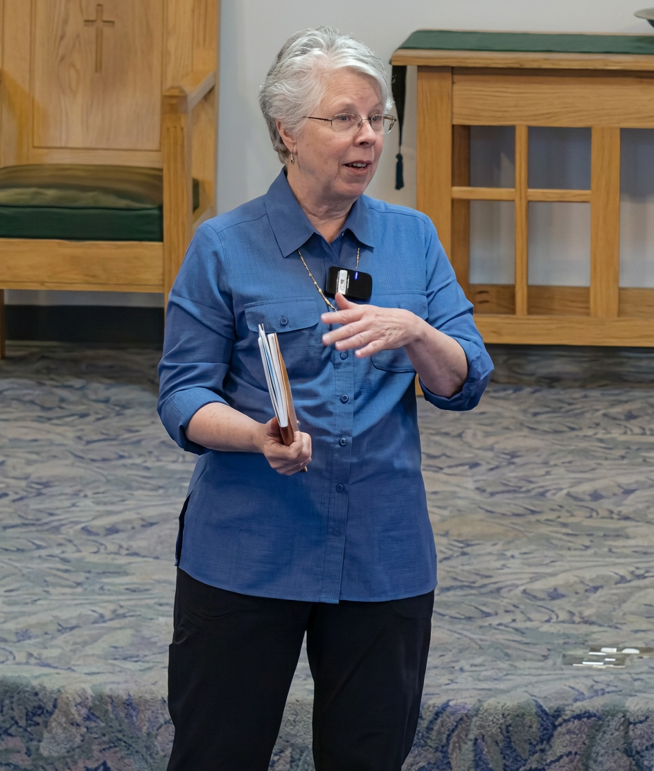 A woman with short gray hair, wearing glasses and a blue shirt, is standing and speaking in a room with wooden furniture. She is gesturing with one hand while holding papers in the other.