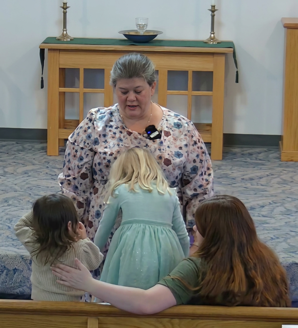 A woman in a floral blouse interacts with two young girls in a church setting. The girls are focused on an activity, and another woman is seated in the background, gently touching one of the girls.