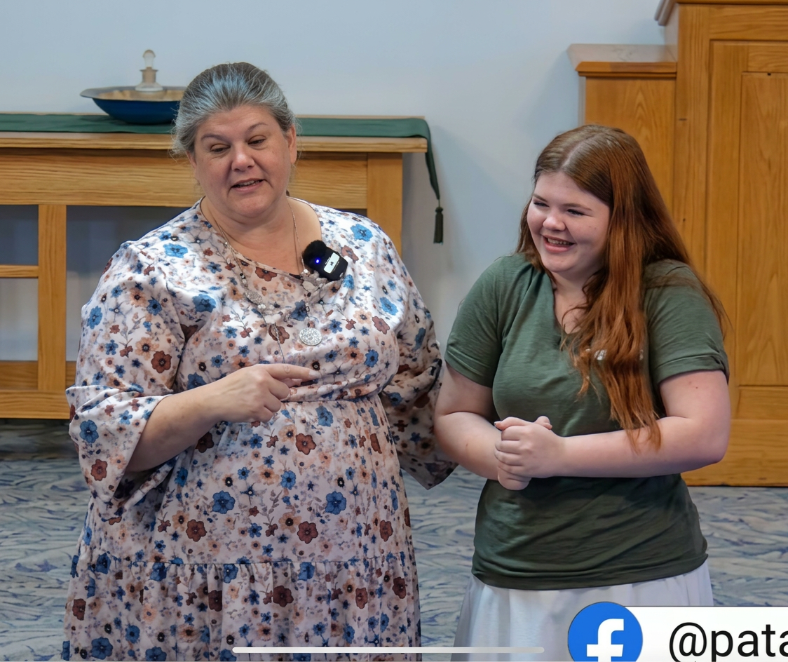 A woman in a floral dress speaks animatedly, pointing while sharing a moment with a smiling young girl dressed in a green shirt. They are both engaged and appear to be in a friendly conversation, standing in a well-lit room with wooden furniture.