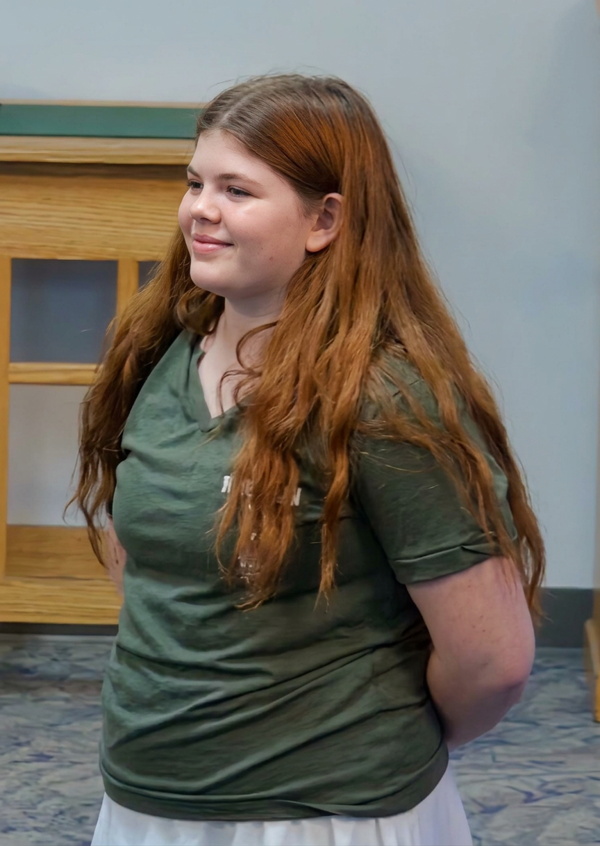 Profile view of a young girl with long red hair, wearing a green shirt, smiling and standing with her hands behind her back.